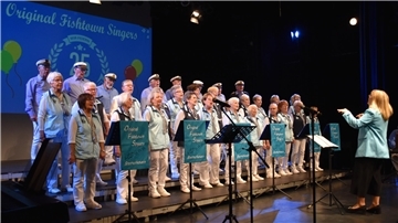 Der Chor "Original Fishtown Singers" aus Bremerhaven steht in maritimer Kleidung auf einer Bühne und singt unter der Leitung einer Dirigentin. Im Hintergrund zeigt eine Leinwand das 25-jährige Jubiläum des Chores.