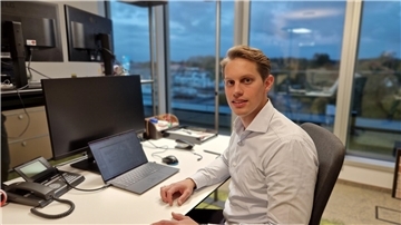 Ein Mann sitzt an einem Schreibtisch, auf dem ein PC steht, und lächelt in die Kamera.