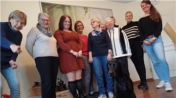 Eine Gruppe von überwiegend älteren Frauen lächelt in die Kamera, vorne ein schwarzer Hund.  