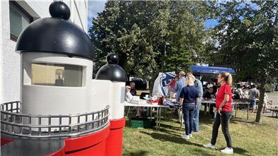 Ein detailreiches Modell eines Leuchtturms steht im Vordergrund. Dahinter findet bei sonnigem Wetter ein Fest mit Besuchern an Biertischen und Infoständen statt. Menschen unterhalten sich, im Hintergrund ist ein mobiler Kaffee- oder Essensstand zu erkennen.