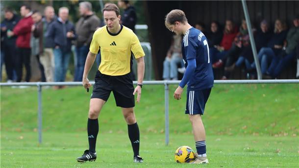 Auf einem Fußballplatz: Ein Mann mit einer Pfeife in der Hand spricht mit einem anderen Mann, zu dessen Füßen ein Fußball liegt. Im Hintergrund stehen Zuschauer.