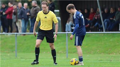 Auf einem Fußballplatz: Ein Mann mit einer Pfeife in der Hand spricht mit einem anderen Mann, zu dessen Füßen ein Fußball liegt. Im Hintergrund stehen Zuschauer.