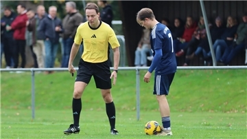 Auf einem Fußballplatz: Ein Mann mit einer Pfeife in der Hand spricht mit einem anderen Mann, zu dessen Füßen ein Fußball liegt. Im Hintergrund stehen Zuschauer.