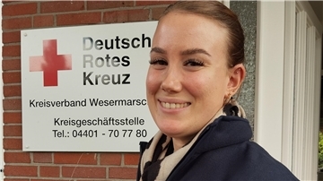 Eine junge Frau vor dem Schild des DRK Wesermarsch. Altenpflegerin Chariece Hübler ist im DRK-Kreisverband für die Pflegeberatung zuständig.