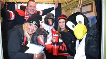 Eishockey-Fans sitzen in einem Zug und strahlen mit Pinguins-Maskottchen in die Kamera.