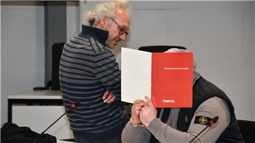 Anwalt sitzt neben einem Angeklagten, der sich eine Mappe vor das Gesicht hält.