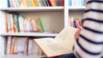 Ein Schüler liest ein Buch, im Hintergrund ist ein Regal mit vielen Büchern zu sehen.