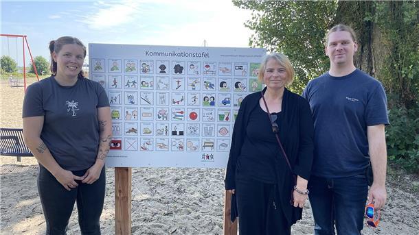 Drei Personen stehen am Strand vor einer Tafel mit Piktogrammen.