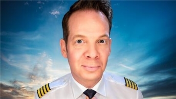 Ein Mann in Pilotenuniform mit vier goldenen Streifen auf den Schulterklappen blickt lächelnd in die Kamera. Im Hintergrund ist ein Himmel mit Wolken und Sonnenuntergang zu sehen.