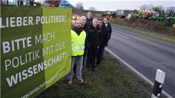 Männer am Straßenrand mit Plakat, Trecker im Hintergrund.