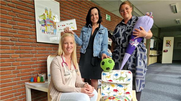 Drei Frauen sitzen und stehen an einem kleinen Tisch mit Spielzeug und einem Kinderstuhl mit Dinosaurier-Muster