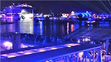 Nachtaufnahme eines Hafens mit einem großen, beleuchteten Kreuzfahrtschiff im Hintergrund, das das charakteristische Design von Augen und Lippen auf der Schiffswand trägt. Der Hafen ist in blauem Licht illuminiert, mit zahlreichen Menschen, die das Szenario von einer Aussichtsplattform und einer unteren Ebene beobachten.