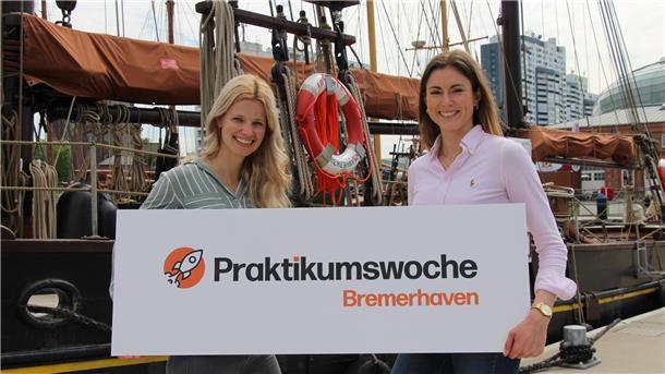 Zwei junge Frauen, Mitarbeiterinnen aus dem Standortmarketing der BIS Wirtschaftsförderung, halten vor der Kulisse des Neuen Hafens in Bremerhaven, ein Banner mit der Aufschrift "Praktikumswoche Bremerhaven"