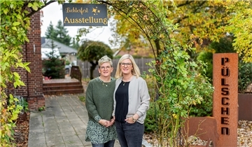 Zwei Frauen vor einem grün drapierten Eingangsbereich. Rechts von ihnen steht auf einem Schild "Pülschen", über ihnen hängt eine Tafel mit der Aufschrift "Lichterfest, Ausstellung".
