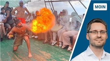 Ein Mann, der auf dem Deck eines Schiffes kniet. Er pustet eine Flüssigkeit aus, mit der er einen Feuerball erzeugt. Publikum sitzt um ihn herum.