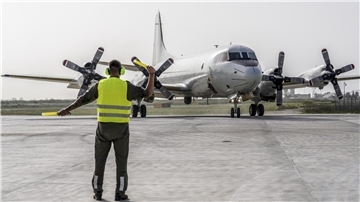 Ein Soldat mit Gehörschutz und gelber Warnweste weist ein viermotoriges Propellerflugzeug vom Typ P-3C Orion auf einem Flugfeld ein. Das Flugzeug befindet sich im Rollvorgang.