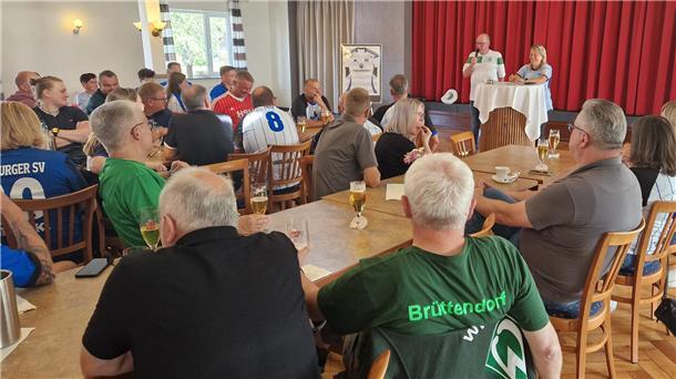 Top, die Wette gilt: Traditionelle Fußballwetten erleben ein Comeback Auf einem Festsaal sitzen Menschen an langen Tischen. Vor der Bühne steht ein Mann mit einem Mikrofon in der Hand und eine Frau an einem Stehtisch.