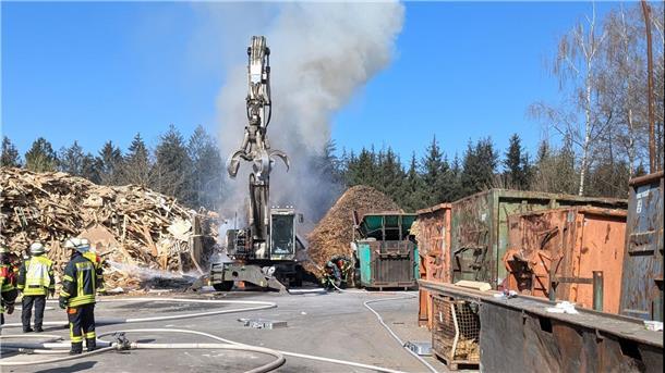 Ein Bagger qualmt. Feuerwehrleute stehen zur Löschung bereit.