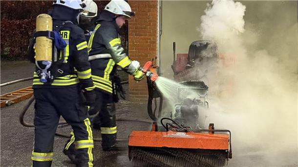 Rasentraktor gerät in Brand: 10.000 Euro Schaden in Elsdorf Drei Männer in schwarz- und signalgelben Anzügen halten einen Wasserstrahl auf einen brennenden Aufsitzrasenmäher.