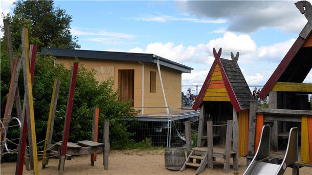 Auf einem sandigen Bogen stehen Spielgeräte für Kinder. Ein kleines Gebäude befindet sich im Bau. 