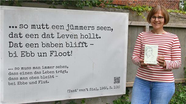 Barbara Müller steht neben einem Plakat. Darauf ist ein niederdeutsches Zitat zu sehen, das ins Hochdeutsche übersetzt wurde.