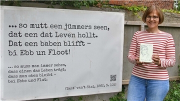 Barbara Müller steht neben einem Plakat. Darauf ist ein niederdeutsches Zitat zu sehen, das ins Hochdeutsche übersetzt wurde.