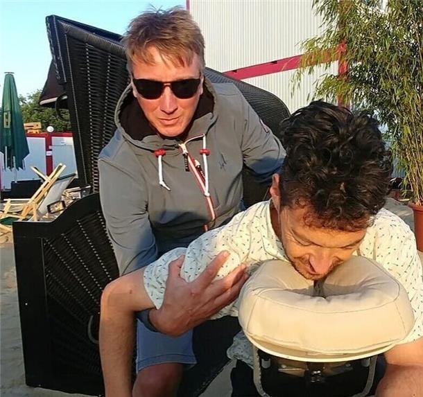 Ein Masseur behandelt einen Mann in einer Massageliege inmitten von Sand und Strandkörben