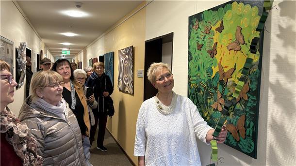 Susanne Schier zeigt ihre Werke in der Kleinen Galerie im Loxstedter Rathaus Eine Gruppe von Menschen steht in einem Flur und betrachtet ein Kunstwerk an der Wand. Die Künstlerin, in weiß gekleidet, lächelt und zeigt auf das Bild, das aus grünen und braunen Elementen besteht und Naturmotive darstellt.