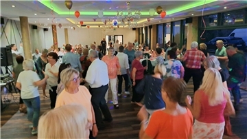 Zahlreiche Menschen tanzen und feiern in einem festlich geschmückten Saal mit Ballons und bunter Beleuchtung; im Hintergrund sitzen Gäste an runden Tischen.