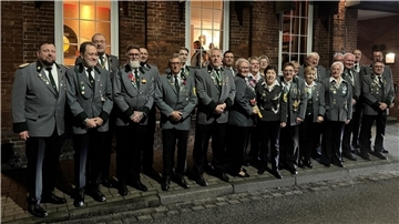 Eine Gruppe von Personen in grauen Uniformjacken mit Abzeichen steht in einer Reihe vor einem Backsteingebäude. Die Gruppe besteht aus Männern und Frauen unterschiedlichen Alters, die lächeln und für ein Foto posieren.