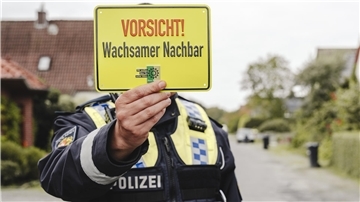 Ein Polizeibeamter hält ein gelbes Schild mit der Aufschrift „Vorsicht! Wachsamer Nachbar“ in die Kamera. Im Hintergrund ist eine ruhige Wohnstraße zu sehen.