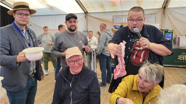 Drei Herren stehen beisammen. Einer hält einen Schweine-Schwanz in der Hand und hält diesen einer Dame vor die Nase. 