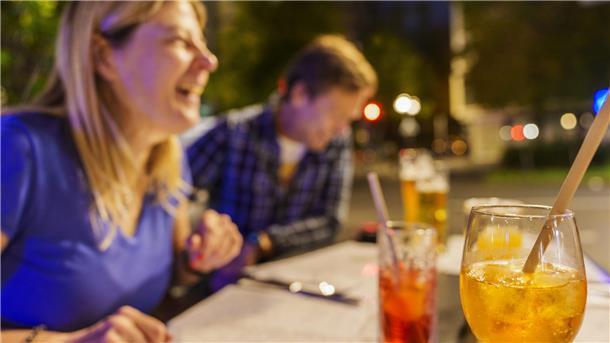 Eine lachende Frau und ein lachender Mann sitzen mit Cocktails an einem Tisch. 