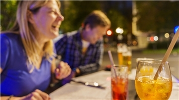 Eine lachende Frau und ein lachender Mann sitzen mit Cocktails an einem Tisch. 