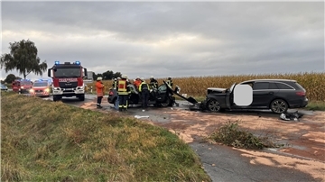 Mehrere Einsatzfahrzeuge von Feuerwehr, Rettungsdienst und Polizei stehen auf einer von einem Verkehrsunfall verwüsteten Landstraße. Im Vordergrund sind zwei durch die Kollision schwer beschädigte Pkw zu sehen.