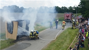 Zwei kniende Feuerwehrleute richten Wasser aus einem Löschschlauch auf einen nur noch rauchenden Holzverschlag. In sicherer Entfernung beobachtet Publikum die Löschung dieses simulierten Zimmerbrands.