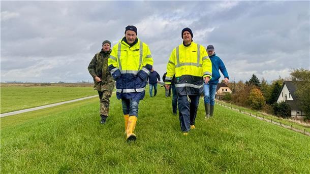 Mehrere Personen in wetterfester Kleidung und Warnjacken gehen bei windigem Wetter über einen grünen Deich. Im Hintergrund sind Felder, Häuser und ein grauer Himmel zu sehen.