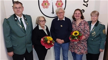 Fünf Personen stehen vor einem Wandwappen, zwei Männer und drei Frauen. Zwei Frauen und ein Mann in der Mitte halten Blumensträuße, die äußeren Personen tragen grüne Schützenuniformen mit Abzeichen.