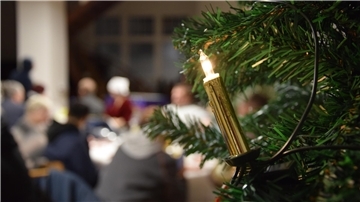 Eine Kerze brennt am Weihnachtsbaum. Im Hintergrund sitzen Wohnungslose beim Weihnachtsessen zusammen. 