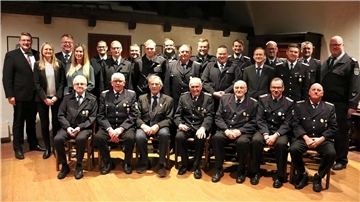 Eine größere Gruppe von Personen posiert für ein Gruppenfoto in einem Innenraum. In der vorderen Reihe sitzen ältere Herren in Feuerwehruniformen, einige mit Auszeichnungen auf der Brust. Hinter ihnen stehen weitere Männer und Frauen, teils in Uniform, teils in formeller Kleidung. 