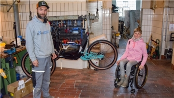 Ein Mann steht in einer Werkstatt, hinter ihm hängt ein Handbike am Haken, neben ihm sitzt eine Rollstuhlfahrerin.