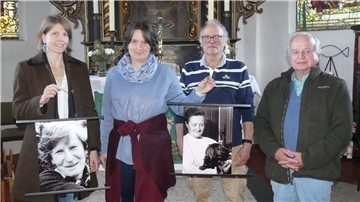 Zwei Frauen und zwei Männer stehen vor dem Altar einer Kirche. Sie halten zwei Schwarz-Weiß-Aufnahmen in Händen, die Porträtfotos zweier Frauen zeigen.