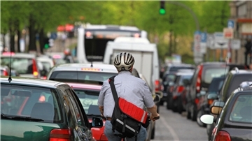 **ALT-Text:**
Ein Radfahrer mit Helm fährt zwischen dichtem Autoverkehr in einer städtischen Straße, umgeben von Autos, Bussen und Ampeln bei grünem Licht.