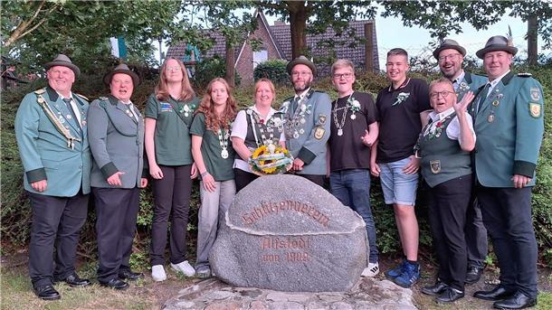 Mitglieder des Schützenvereins Alfstedt, darunter die frisch gekrönten Majestäten, stehen in Uniform und Freizeitkleidung fröhlich lächelnd um einen Gedenkstein mit der Inschrift „Schützenverein Alfstedt von 1909“. Die Gruppe posiert im Grünen vor einer Hecke.