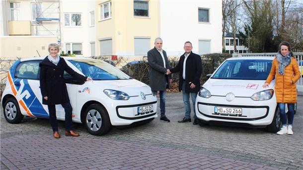 Das Foto zeigt vier Menschen und zwei Autos. Die beiden Herren geben sich die Hände.