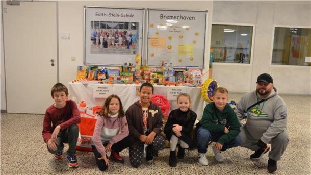 Klassensprecher der 5. Klassen der Edith-Stein-Schule und Silvio Späte vom Verein "Rückenwind für Leher Kinder" knien vor dem Gabentisch mit den gespendeten Lebensmitteln, Büchern, Spielzeug und Bekleidung