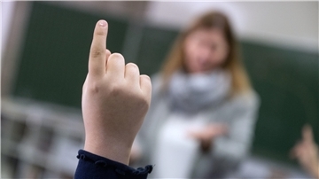 Eine Hand mit ausgestrecktem Finger. Im Hintergrund eine Lehrerin.  