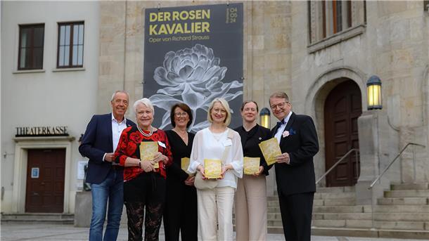 vier Frauen und zwei Männer stehen vor dem Eingang des Stadttheaters Bremerhaven, an dessen Fassade ein großes Plakat hängt mit der Aufschrift "Der Rosenkavalier – Oper von Richard Strauss"