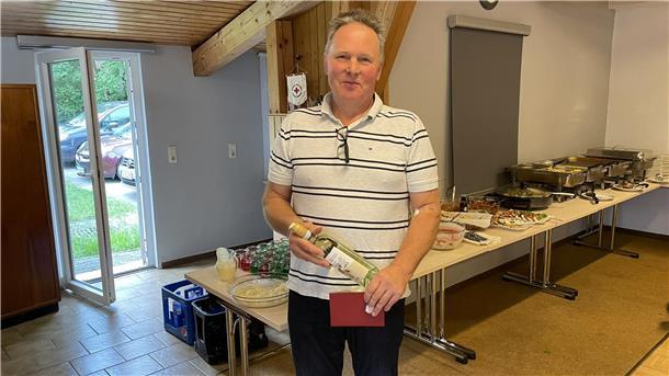 Ein Mann hält ein Präsent in Händen. Hinter ihm ist ein großes Buffet aufgebaut. 