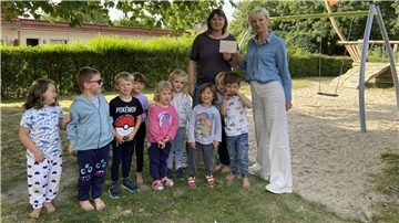 Eine Frau reicht einer anderen Frau im Beisein von Kindern einen Scheck. 